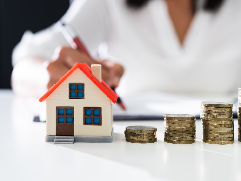 Moedas empilhadas ao lado de miniatura de casa e pessoa escrevendo, sugerindo planejamento financeiro ao investir em aluguéis.