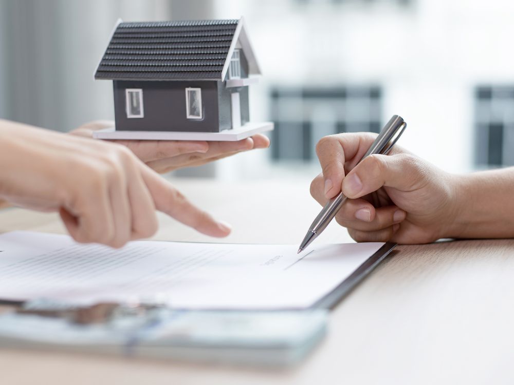 Mão assinando contrato com maquete de casa na mesa, representando segurança ao investir em aluguéis como estratégia de renda passiva mensal.