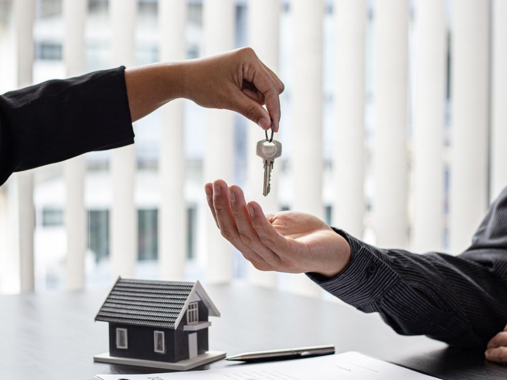 Mão entregando chave em mesa com maquete de imóvel e contrato, representando negociação para alugar uma casa ou apartamento com segurança.