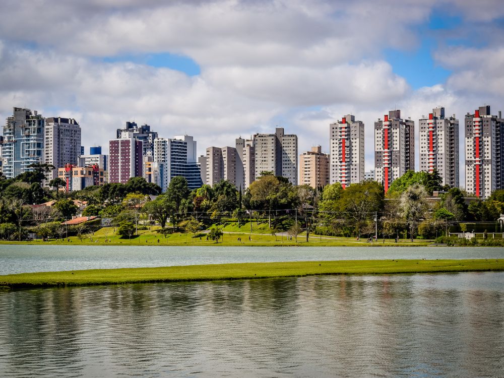 Vista panorâmica de Curitiba com prédios modernos ao fundo e um lago cercado por área verde, destacando a qualidade de vida da cidade e o IDH de Curitiba.