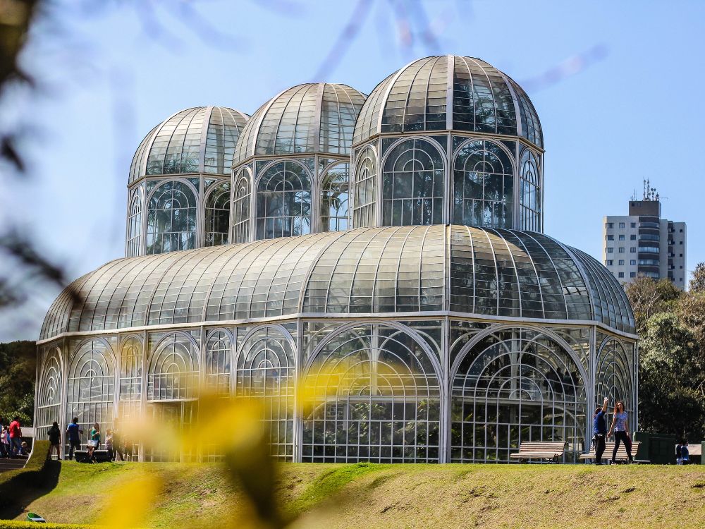 Detalhes do Jardim Botânico de Curitiba: Estrutura de vidro da estufa com pessoas aproveitando o espaço em um dia ensolarado.