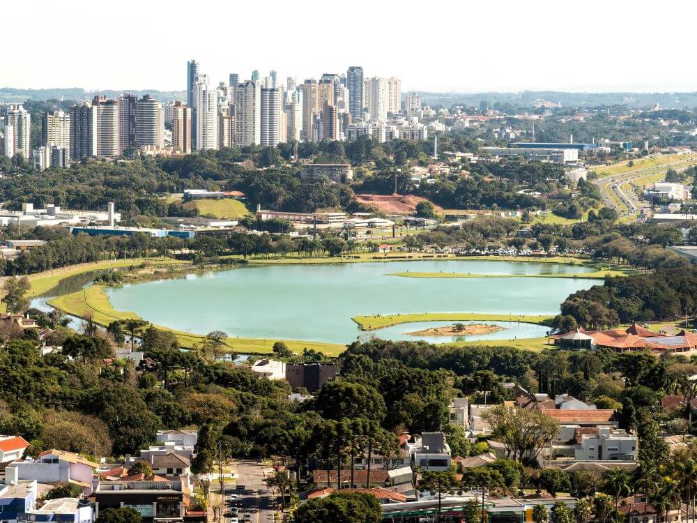 Vista panorâmica de Curitiba com lago, áreas verdes e prédios ao fundo, destacando a diversidade urbana e natural da cidade.