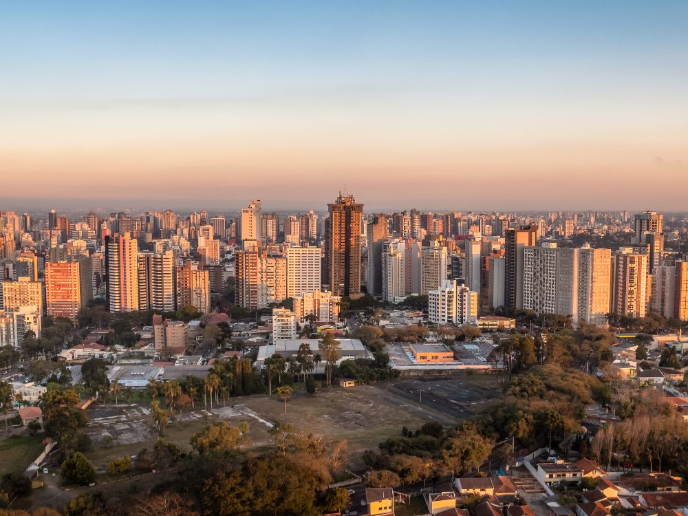 Vista panorâmica de Curitiba ao pôr do sol: Skyline da cidade com prédios altos e áreas arborizadas destacadas pelo céu alaranjado.