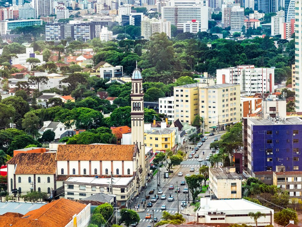 Vista aérea de um bairro central de Curitiba com prédios residenciais, igreja histórica e ruas arborizadas, destacando a qualidade de vida urbana.