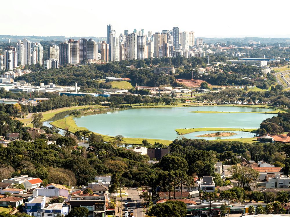 Vista aérea do Parque Barigui em Curitiba, com o lago e áreas verdes cercados por prédios modernos e infraestrutura urbana. Morar em Curitiba é caro ou barato?
