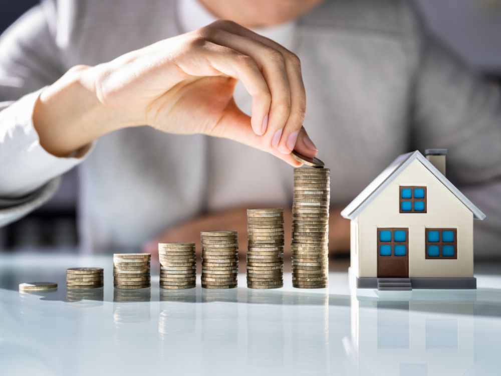 Pessoa empilhando moedas ao lado de uma maquete de casa, representando o crescimento do capital com investimento no mercado imobiliário.