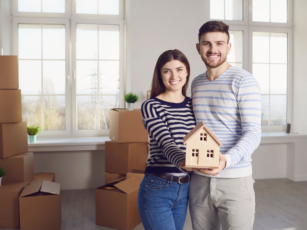Casal sorridente segurando uma maquete de casa entre caixas de mudança, representando quem decidiu investir em Curitiba com foco em locação.
