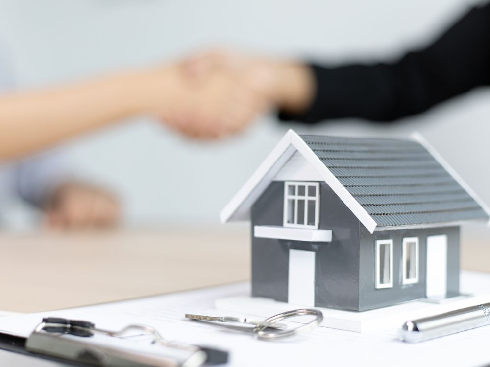 Aperto de mãos entre duas pessoas com maquete de casa e contrato à frente, representando fechamento de negócio em investimento no mercado imobiliário.