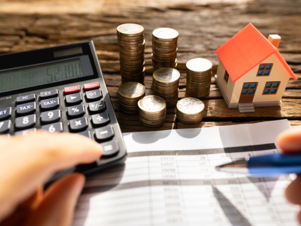 Calculadora, moedas e miniatura de casa sobre planilha, representando como calcular rentabilidade de aluguel com base em custos e retorno anual.
