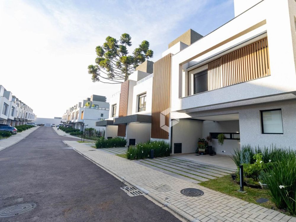 Rua interna de condomínio residencial moderno com casas geminadas de dois andares, fachadas com design contemporâneo e jardim frontal bem cuidado.