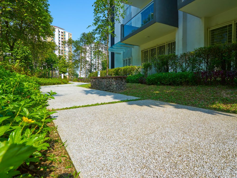 Área externa de condomínio com calçada de concreto, jardins arborizados e plantas ornamentais, ao lado de prédio residencial moderno com varandas envidraçadas.