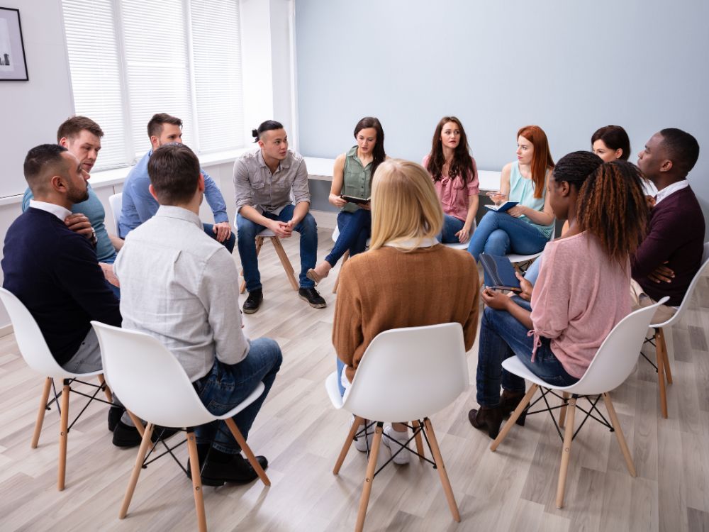 Grupo de pessoas reunidas em círculo em uma sala iluminada, participando de reunião ou dinâmica em grupo, sentadas em cadeiras brancas sobre piso laminado claro. Discutindo se taxa de condomínio inclui água e luz