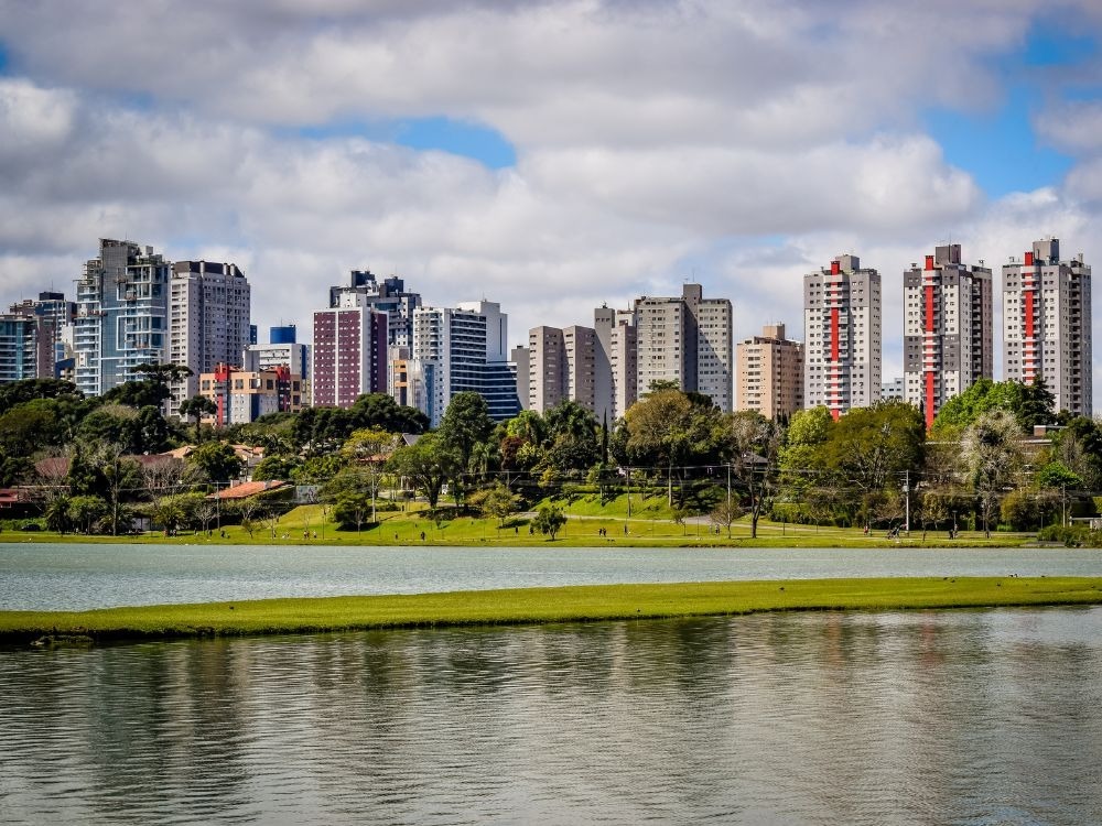 Vista aérea do centro de Curitiba, mostrando vários edifícios residenciais modernos e um grande parque verde. A imagem promove a cidade como uma das Melhores cidades para investir em imóveis.