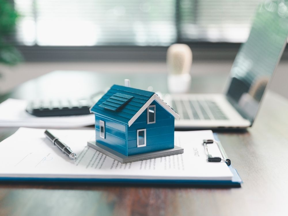 Maquete de casa azul sobre contrato imobiliário com notebook ao fundo, simbolizando a formalização e consulta do CPF dos imóveis.