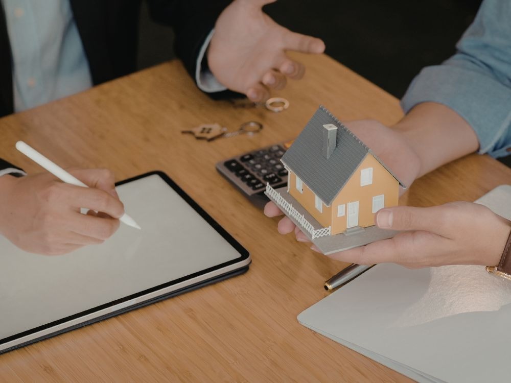 Corretor apresentando maquete de casa enquanto cliente utiliza tablet para análise de dados, representando a importância do CPF dos imóveis na organização documental.