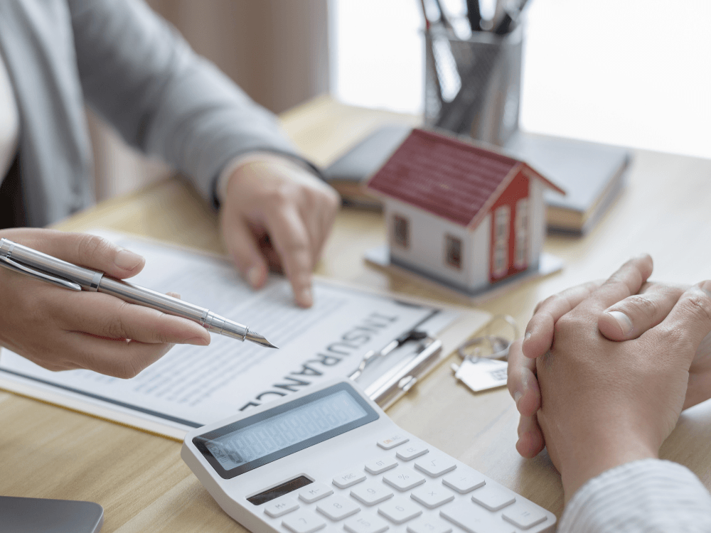 Atendimento com contrato, calculadora e maquete de casa durante conversa sobre despesas e IPTU em imóvel alugado.