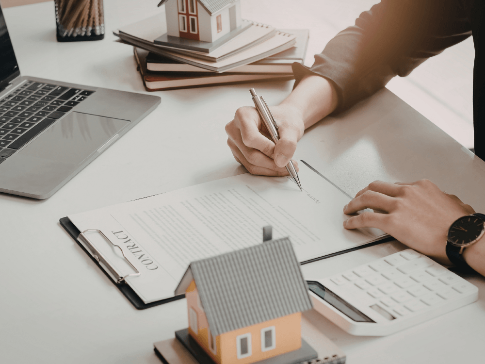 Pessoa assinando contrato de locação com miniatura de casa e calculadora sobre a mesa de trabalho.