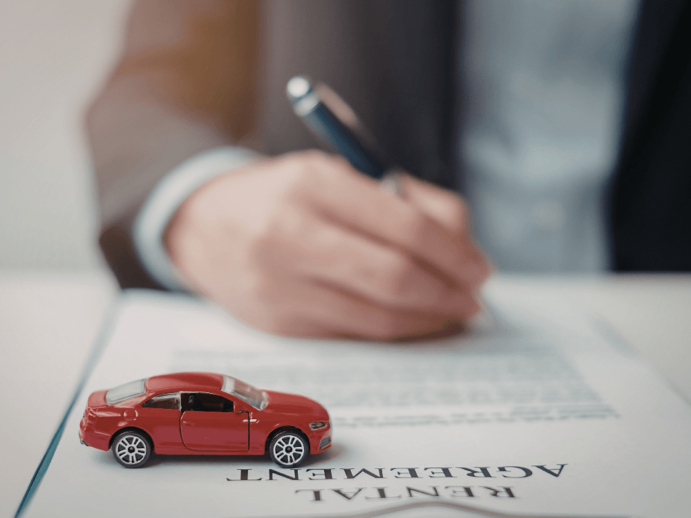 Miniatura de carro sobre documento contratual enquanto uma pessoa assina o termo de garantia da locação.