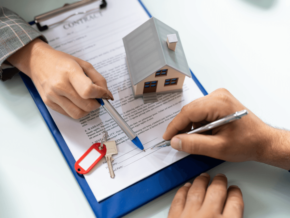 Pessoas assinam um contrato imobiliário ao lado de uma maquete de casa, simbolizando a formalização da venda de imóvel.