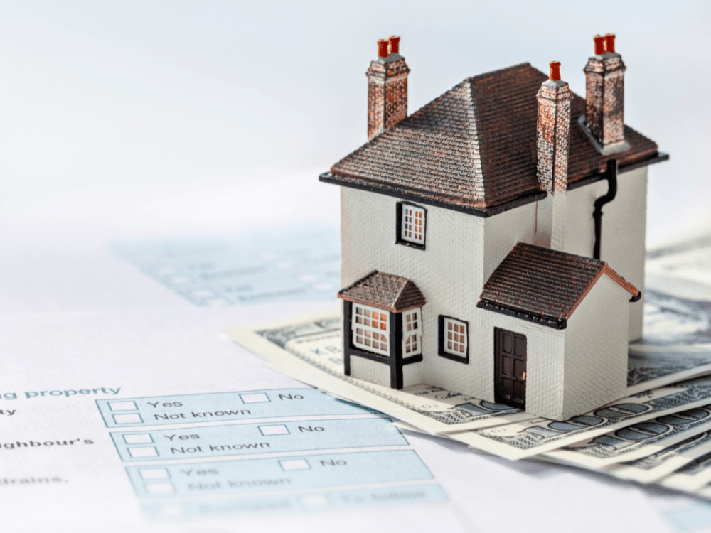 Miniatura de uma casa sobre documentos e dinheiro, ilustrando valores e registros envolvidos na venda de um imóvel. Tudo para explicar se quando se vende um imóvel tem que pagar imposto.