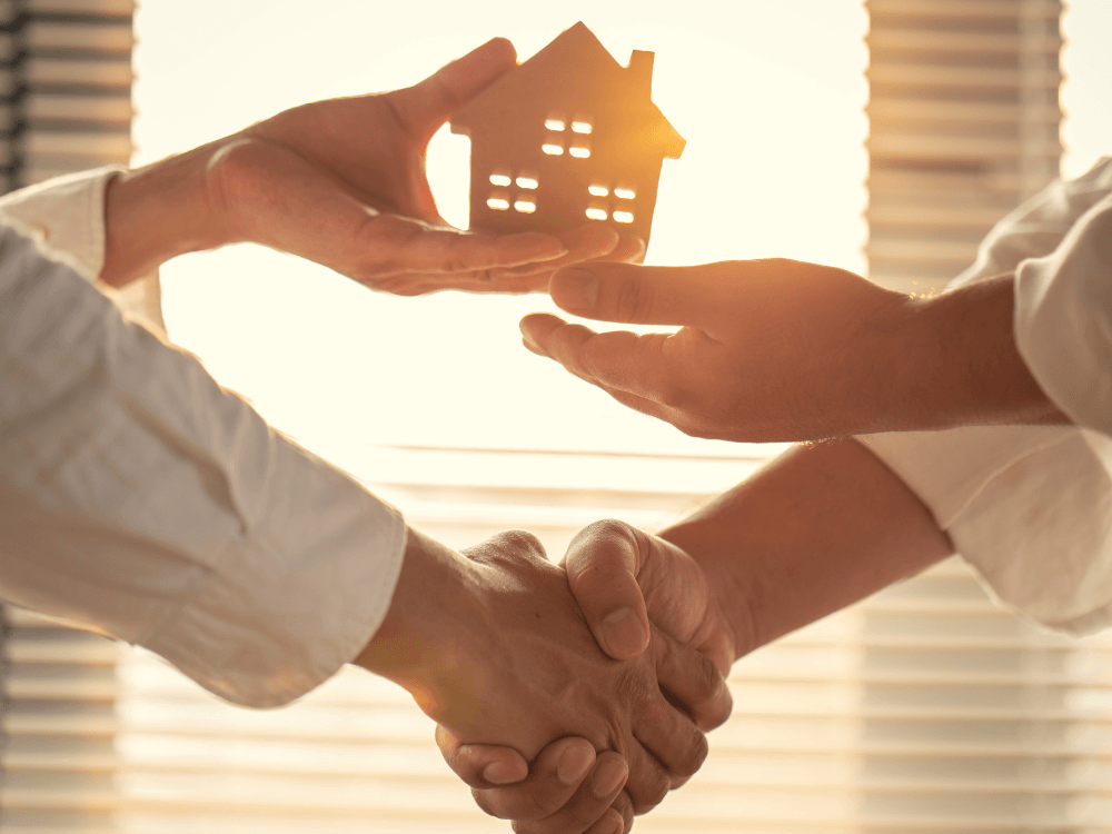 Duas pessoas apertam as mãos enquanto seguram uma maquete de casa, simbolizando uma negociação imobiliária e o cadastro de propriedades.