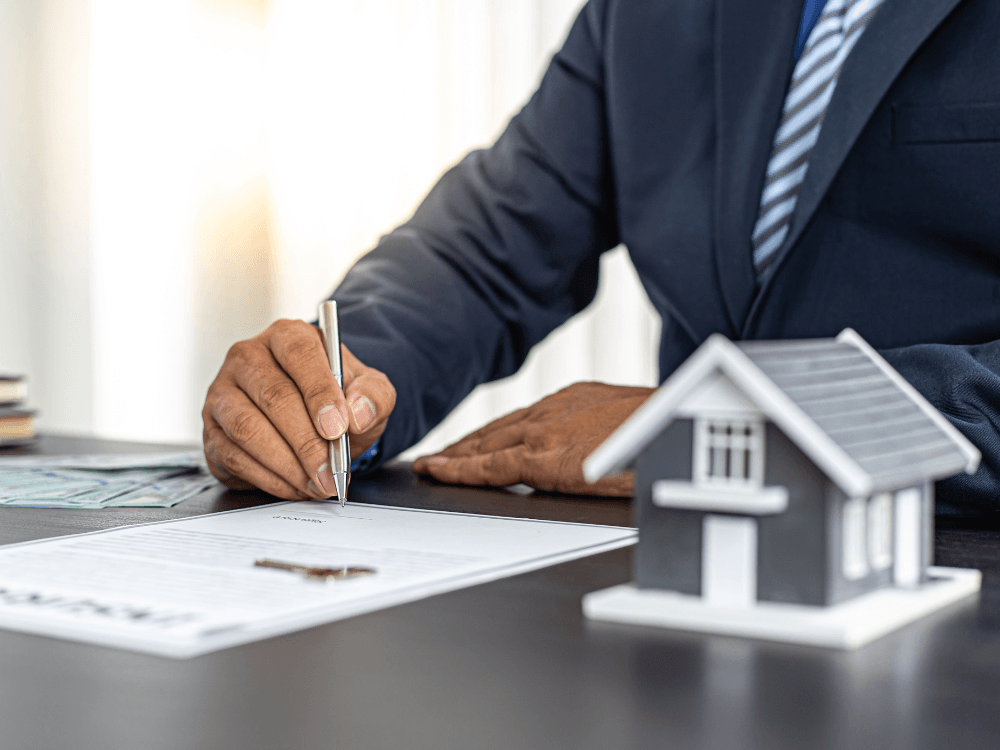 Profissional de terno assina um documento imobiliário sobre uma mesa, com maquete de casa em primeiro plano, representando cadastro de imóveis.