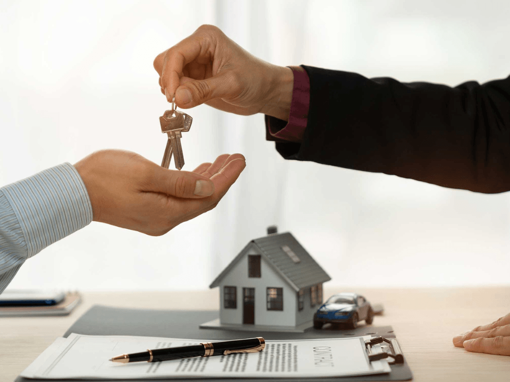 Mãos trocam chaves de uma casa sobre contrato imobiliário, simbolizando a compra e venda de propriedades e seus registros cadastrais.