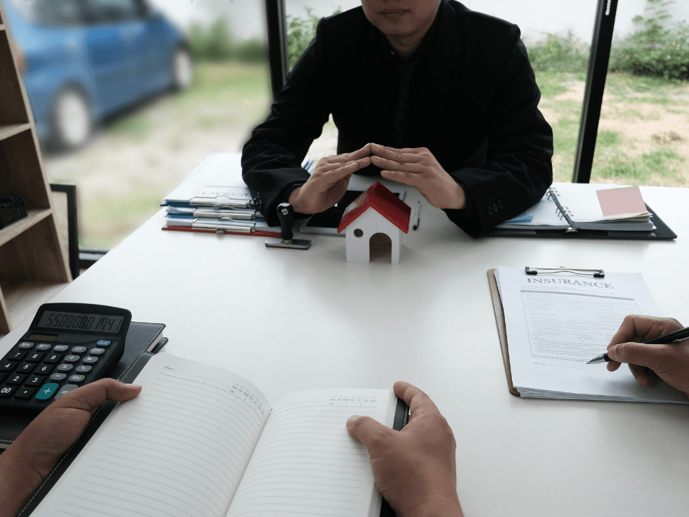 Profissional em escritório analisa documentos e utiliza uma calculadora diante de uma maquete de casa durante uma reunião sobre patrimônio imobiliário.